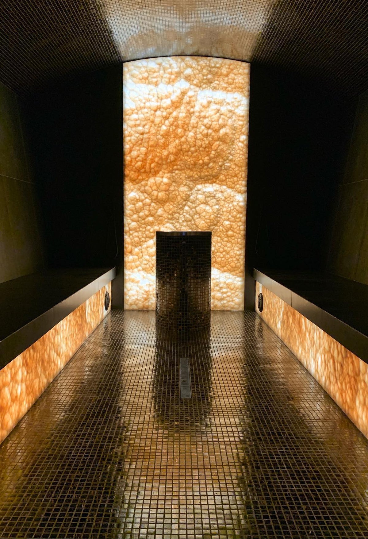 A serene, modern wellness or sauna room interior with a long tiled walkway, glowing orange-brown textured stone walls, and a central illuminated panel.